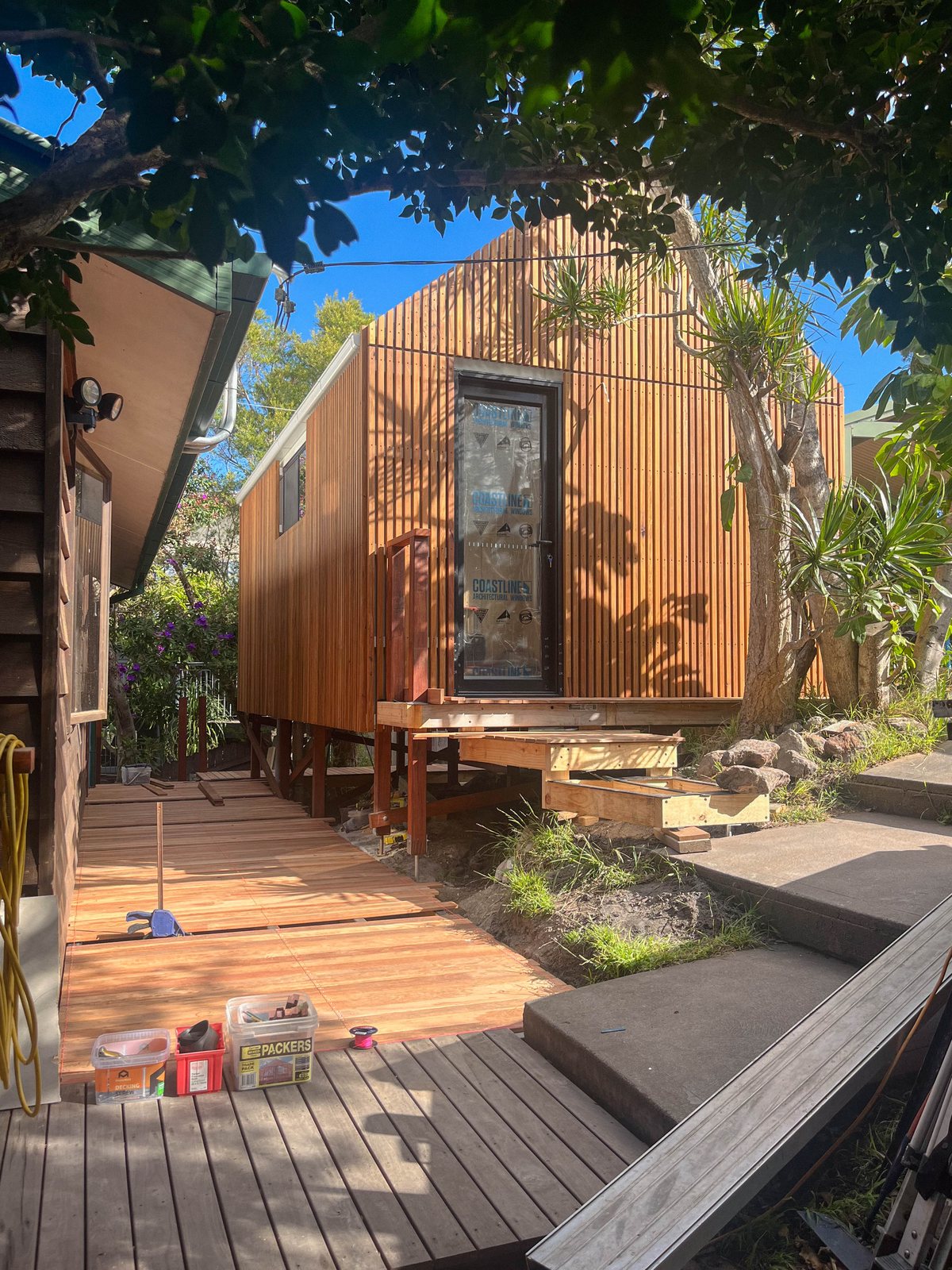 A small building with vertical timber cladding beside decking, tools and exposed framing.
